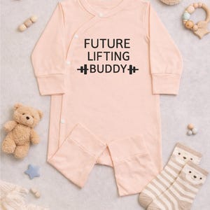 May include: A light peach-colored baby onesie with the text "FUTURE LIFTING BUDDY" in black. The long-sleeved outfit has a wrap-style front with snap closures. A teddy bear, star, and socks with bear faces are also in the image.