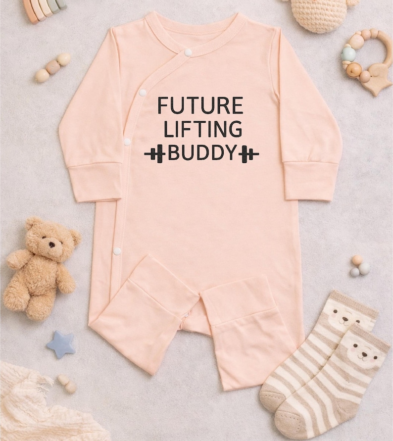 May include: A light peach-colored baby onesie with the text "FUTURE LIFTING BUDDY" in black. The long-sleeved outfit has a wrap-style front with snap closures. A teddy bear, star, and socks with bear faces are also in the image.