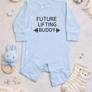 May include: A light blue baby onesie with the text "FUTURE LIFTING BUDDY" in black. The onesie has long sleeves and attached feet. The image also includes baby toys and socks.