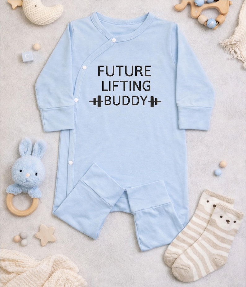 May include: A light blue baby onesie with the text "FUTURE LIFTING BUDDY" in black. The onesie has long sleeves and attached feet. The image also includes baby toys and socks.