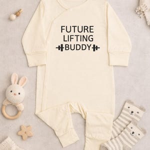 May include: Cream-colored baby onesie with the text "FUTURE LIFTING BUDDY" in black. The onesie has long sleeves and a wrap-style closure. A stuffed bunny, wooden star, and striped bear socks are also in the image.
