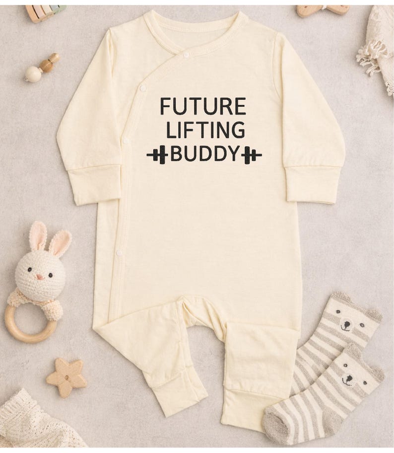 May include: Cream-colored baby onesie with the text "FUTURE LIFTING BUDDY" in black. The onesie has long sleeves and a wrap-style closure. A stuffed bunny, wooden star, and striped bear socks are also in the image.