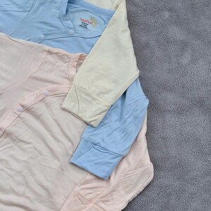May include: Three pastel-colored baby onesies. The onesies are light pink, light blue, and cream. Each onesie has long sleeves and a snap closure. The brand name "SunPod" is printed on the chest of the blue and cream onesies.