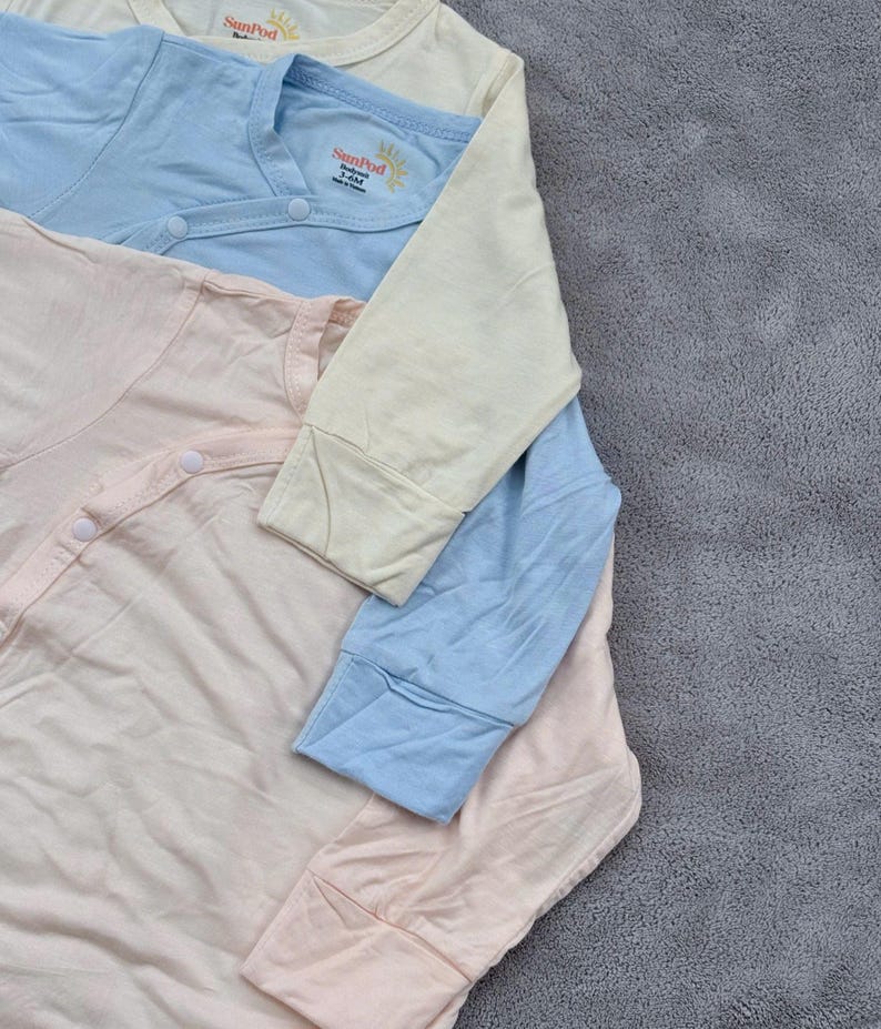 May include: Three pastel-colored baby onesies. The onesies are light pink, light blue, and cream. Each onesie has long sleeves and a snap closure. The brand name "SunPod" is printed on the chest of the blue and cream onesies.