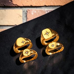 Puede incluir: Cuatro anillos de sello dorados con un diseño de estrella y un pequeño diamante en el centro. Dos anillos tienen una cara ovalada y dos tienen una cara cuadrada. Los anillos se muestran sobre una superficie negra.