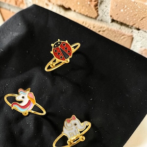 Könnte beinhalten: Drei goldfarbene Ringe mit bunten Emaille-Anhängern. Ein Ring zeigt einen rot-schwarzen Marienkäfer, ein weiterer ein weißes Einhorn mit rosa-blauer Mähne und der dritte einen weißen Elefanten mit einer kleinen rosa Schleife.