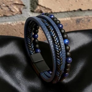 Può includere: Un bracciale a più fili che combina strisce di pelle blu, un cordino intrecciato blu scuro e nero e una fila di perline nero opaco e blu lucido. Il bracciale è fissato con una chiusura magnetica rettangolare in metallo nero opaco.
