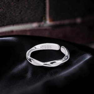 Peut inclure: Une bague ouverte de couleur argentée avec un design torsadé et fluide. La bande intérieure présente le texte gravé S925, indiquant la composition du matériau. La bague a une surface polie et réfléchissante avec une ouverture réglable.