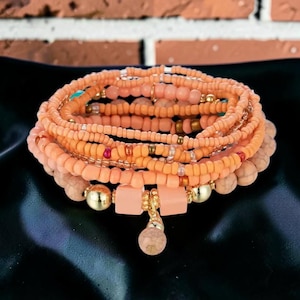 Puede incluir: Una pulsera de múltiples hilos con numerosas cuentas de semillas color melocotón, intercaladas con esferas doradas metálicas, cuentas cuadradas de color melocotón mate y cuentas de piedra beige texturizadas. Una cuenta redonda cuelga al centro.