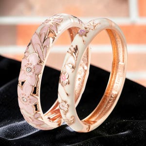Puede incluir: Dos brazaletes de oro rosa con diseños florales. Uno presenta flores rosas con detalles de cristal transparente, mientras que el otro tiene una base de esmalte blanco con patrones florales rosas. Los brazaletes están sobre un fondo negro.