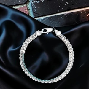 May include: A silver-toned chain bracelet featuring a flat, interlocking herringbone-style link pattern. The bracelet is secured with a polished lobster claw clasp mechanism, showcasing a bright, reflective metallic finish throughout the entire piece.