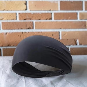 May include: A black headband made from a soft, stretchy fabric. The headband is designed to be worn around the forehead, likely for sports or fashion. The background features a brick wall.