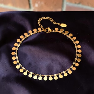 May include: A gold-toned chain anklet featuring a series of small, flat, circular discs suspended along the entire length of the chain. The anklet includes a lobster claw clasp and a small oval tag near the closure that is engraved with the text K18.