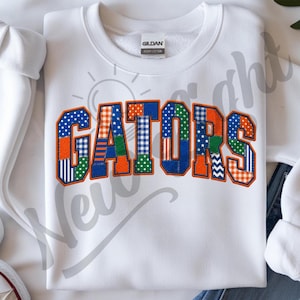Puede incluir: Una sudadera blanca de cuello redondo con la palabra GATORS en letras grandes de estilo bloque. Cada letra está rellena con un mosaico de patrones de tela en azul, naranja y verde, incluyendo lunares, rayas, cuadros y estrellas.
