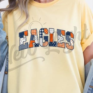 Könnte beinhalten: Ein hellgelbes Kurzarm-T-Shirt mit dem Wort EAGLES auf der Brust. Jeder Buchstabe ist mit einem Patchwork-Muster aus marineblauen und orangefarbenen Stoffen gefüllt, darunter Sterne, Streifen, Karos und Zickzackmuster.
