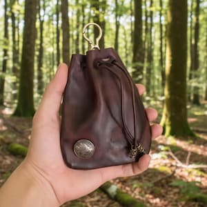 Puede incluir: Una bolsa de cuero marrón oscuro con cierre de cordón y cierre metálico. La bolsa tiene una moneda de plata adjunta en la parte delantera y se sostiene en una mano. El fondo es una escena de bosque borrosa.