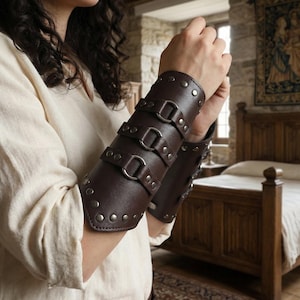 Peut inclure: Bracelets de bras en cuir marron avec rivets et boucles argentés. Les protège-bras ont trois sangles avec des anneaux en D et sont conçus pour protéger l'avant-bras. Le cuir a un aspect légèrement texturé.