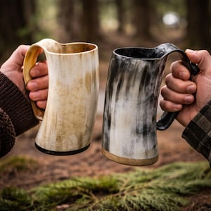 Puede incluir: Dos tazas de cuerno se sostienen frente a un fondo borroso de bosque. Una taza es de color beige claro con un asa curva, la otra es negra y blanca con un asa negra. Ambas tazas tienen un borde oscuro en la base.
