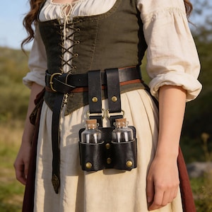 Puede incluir: Cinturón de cuero negro con dos pequeñas botellas de vidrio en fundas de cuero. Las botellas tienen tapones de corcho. El cinturón tiene una hebilla de latón y se usa con un vestido de estilo medieval.