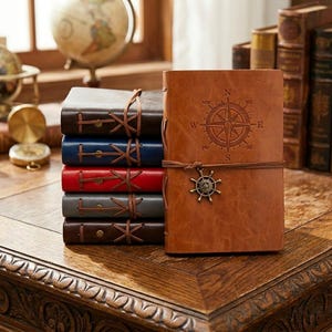 Può includere: Una pila di cinque diari rettangolari con copertine effetto pelle in marrone, blu, rosso, grigio e marrone scuro. Ogni diario presenta una chiusura con cordino avvolgente e un ciondolo in metallo a forma di timone di nave.