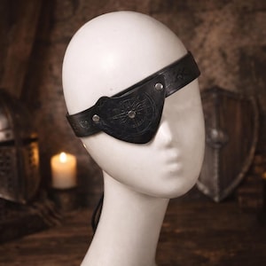 May include: A black leather eye patch with a compass rose design, secured by a strap. The eye patch is displayed on a white mannequin head. The leather has embossed details and is held in place with silver-colored rivets.