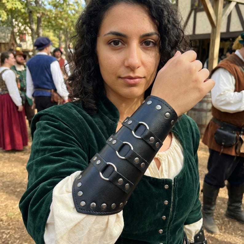 Pu&ograve; includere: Bracciale in pelle nera con rivetti color argento e tre anelli metallici. Il bracciale &egrave; indossato sull'avambraccio e fa parte di un outfit a tema medievale o rinascimentale.