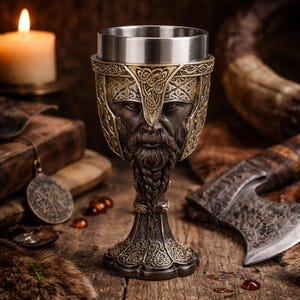 Może przedstawiać: Brązowy kielich ze stalową obręczą. Kielich ma wzór wikinga, z brodatą twarzą i skomplikowanymi detalami hełmu. Kielich stoi na drewnianej powierzchni ze świecą i innymi przedmiotami o tematyce wikingów.