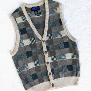 May include: A V-neck sweater vest featuring a patchwork pattern of squares in shades of dark green, grey, and cream. The garment has a button-down front with five brown buttons, a cream-coloured ribbed trim along the neckline, armholes, and hem.