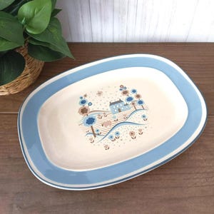 May include: An oval ceramic serving platter with a light blue border and a cream-colored center. The center features a pastoral scene with a house, trees, and flowers in shades of blue, brown, and cream. The platter is a vintage piece.