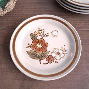 May include: A round ceramic plate with an off-white base and a brown rim. The center features a floral pattern with orange, white, and yellow flowers accompanied by green leaves. The plate has a speckled texture and a smooth, glossy finish.