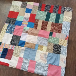 May include: A patchwork quilt constructed from a variety of rectangular and square fabric pieces. The quilt features a diverse array of colors including red, blue, pink, green, and beige, alongside various floral, polka dot, and striped patterns.