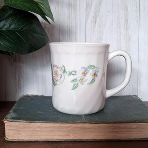 May include: White ceramic mug with a floral design featuring pink and blue flowers. The mug has a curved handle and is set against a backdrop of a weathered book and a light-colored wooden surface. The mug is approximately 4 inches tall.