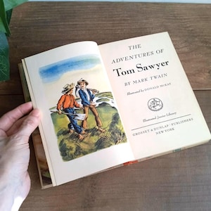 Könnte beinhalten: Ein offenes Buch mit dem Titel "The Adventures of Tom Sawyer" von Mark Twain, illustriert von Donald McKay. Die Illustration zeigt zwei Jungen mit Werkzeugen. Das Buch stammt aus der Illustrated Junior Library, herausgegeben von Grosset & Dunlap, New York.