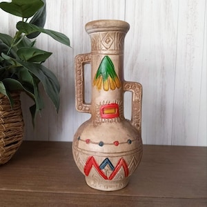 May include: A beige ceramic vase with a textured surface and two handles. The vase features colorful geometric designs in red, blue, and green. The vase has a bulbous body and a cylindrical neck.