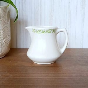 May include: A white ceramic creamer with a green floral design around the top. The creamer has a small handle and a rounded body.