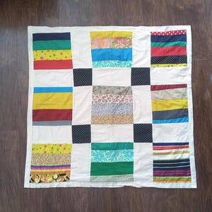 May include: A patchwork quilt featuring a grid of white fabric squares interspersed with blocks of multi-colored fabric strips. The colorful sections include various patterns such as polka dots, floral prints, stripes, and solid colors in red, blue, green, and yellow.