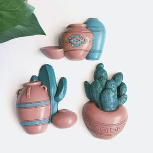 May include: Three decorative wall art pieces featuring Southwestern designs. The pieces include a pink vase with a blue geometric pattern, a blue vase, a small pink bowl, a pink vase with blue stripes, a turquoise cactus, a small pink pot, and a cactus in a pink pot.