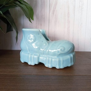 Puede incluir: Una maceta de cerámica azul claro con forma de zapato. La maceta tiene una suela gruesa y detalles decorativos. El zapato es de color azul claro liso y se asienta sobre una superficie marrón oscuro. Una planta verde es visible en el fondo.