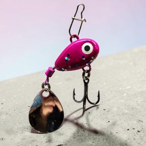 May include: A fishing lure featuring a bright pink teardrop-shaped head with blue glitter accents and a white eye. The lure includes a silver-colored metal spinner blade, a sharp treble hook, and a metal snap swivel attached to the top for connection.