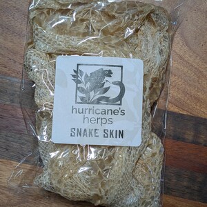 May include: A package of snake skin. The snake skin is a light tan color and is packaged in a clear plastic bag. The label reads "hurricane's herps SNAKE SKIN" with a logo of a snake and leaves.