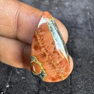 May include: A teardrop-shaped gemstone with a polished surface. The stone features a blend of reddish-brown, black, and green hues, with intricate patterns and textures. The gemstone is held between fingers.