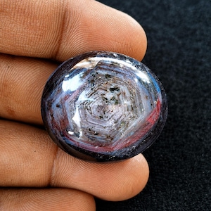 Può includere: Una gemma di zaffiro lucidata di forma ovale caratterizzata da un distinto motivo di zonizzazione del colore esagonale in tonalità blu profondo, rosso-viola e bianco, con inclusioni minerali scure visibili sulla superficie liscia e bombata.