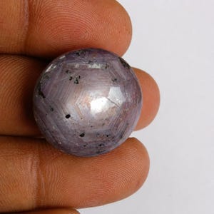 Könnte beinhalten: Ein runder, im Cabochon-Schliff gehaltener Sternrubin-Edelstein in einem durchscheinenden, staubigen Violettton. Die Oberfläche zeigt ein ausgeprägtes hexagonales Wachstumsmuster und verstreute dunkle Mineraleinschlüsse mit einem hellen weißen Lichtreflex in der Mitte.