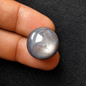 Op de afbeelding: Een ronde, cabochon geslepen edelsteen met een glad, gepolijst oppervlak. De steen heeft een donker, metaalachtig grijze kleur met subtiele blauwe ondertonen en een reflecterende glans over de bolle bovenkant, met zichtbare natuurlijke insluitingen.