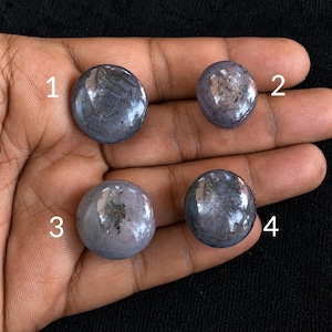 Könnte beinhalten: Vier runde, polierte Cabochon-Edelsteine mit einem gräulich-blauen Farbton und sichtbaren sternartigen Einschlüssen. Jeder Stein ist mit einer weißen Zahl von eins bis vier markiert und in einem Raster auf dunklem Hintergrund angeordnet.