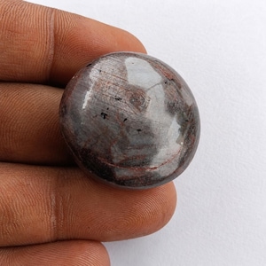 Könnte beinhalten: Ein runder, polierter Cabochon-Edelstein mit einer Mischung aus tiefen rotbraunen und dunkelgrauen Tönen. Die Oberfläche hat ein glattes, reflektierendes Finish mit sichtbaren natürlichen Einschlüssen und subtilen linearen Mustern.