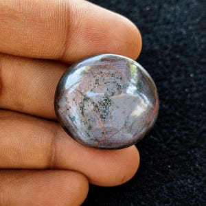 Könnte beinhalten: Ein runder, polierter Cabochon-Edelstein mit einer dunklen, gesprenkelten Oberfläche in tiefen rotbraunen und anthrazitfarbenen Tönen. Der Stein zeigt eine glatte, reflektierende Oberfläche mit sichtbaren Einschlüssen und feinen grünen Flecken.
