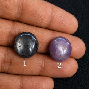 Peut inclure: Deux pierres précieuses cabochons rondes. La pierre 1 est d'un gris métallique foncé avec une surface lisse et polie. La pierre 2 est d'un violet clair translucide avec un motif en forme d'étoile visible. Les pierres portent les numéros 1 et 2.