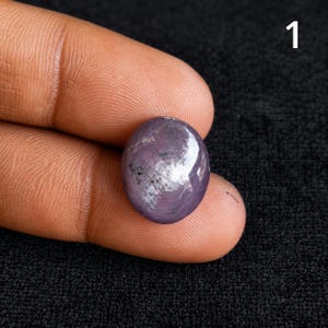 Peut inclure: Une pierre précieuse cabochon de forme ovale avec une teinte violette et grise profonde, présentant un reflet lumineux en forme d'étoile sur sa surface polie. La pierre montre des inclusions naturelles et un sommet lisse et arrondi, marqué du chiffre 1.