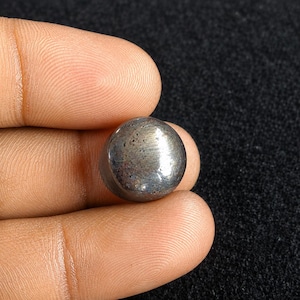 Puede incluir: Un pequeño cabujón circular de aspecto metálico con una superficie lisa y pulida. El objeto presenta un color gris plomo oscuro con sutiles inclusiones moteadas y un brillo reflectante en su parte superior redondeada y abovedada.